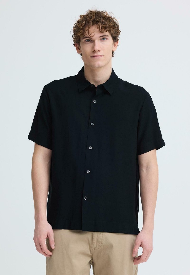 Jeune homme aux cheveux bouclés portant une chemise noire à manches courtes avec des boutons et un pantalon beige, debout devant un fond uni clair.