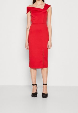 Femme portant une robe midi rouge ajustée à épaules dénudées avec une fente frontale et des talons noirs à bride cheville, debout devant un fond blanc uni.