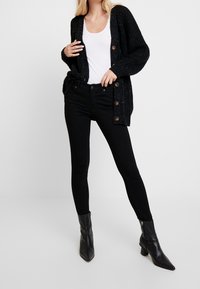 Cardigan en tricot noir avec de gros boutons, porté par-dessus un débardeur blanc et un jean skinny noir. Une paire de bottines noires à bouts pointus complète le look.