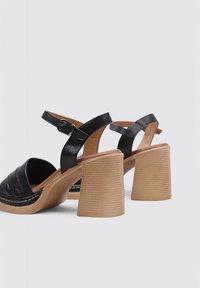 Sandalias de tacón de cuero negro con un diseño de tiras cruzadas, que incluyen una hebilla. El tacón grueso y estriado es de un color marrón claro.