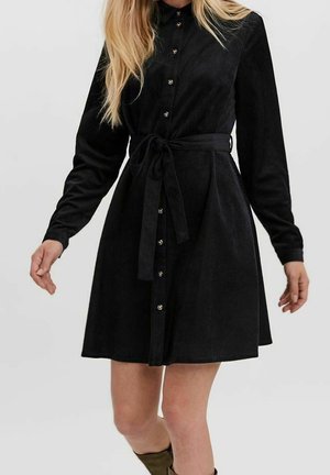 Vestito nero in velluto a coste con maniche lunghe, chiusura frontale con bottoni e cintura aderente in vita. Texture distintiva con coste verticali e gonna svasata.