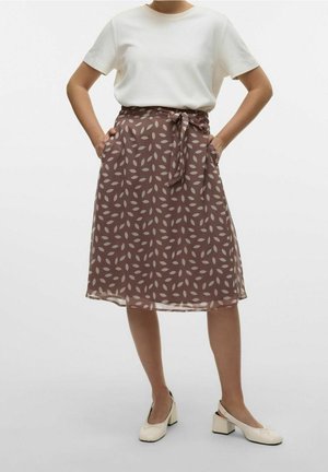 Vrouw draagt een wit shirt met korte mouwen, een knielange bruine rok met licht bladpatroon en een strikceintuur, en crèmekleurige schoenen met blokhak.