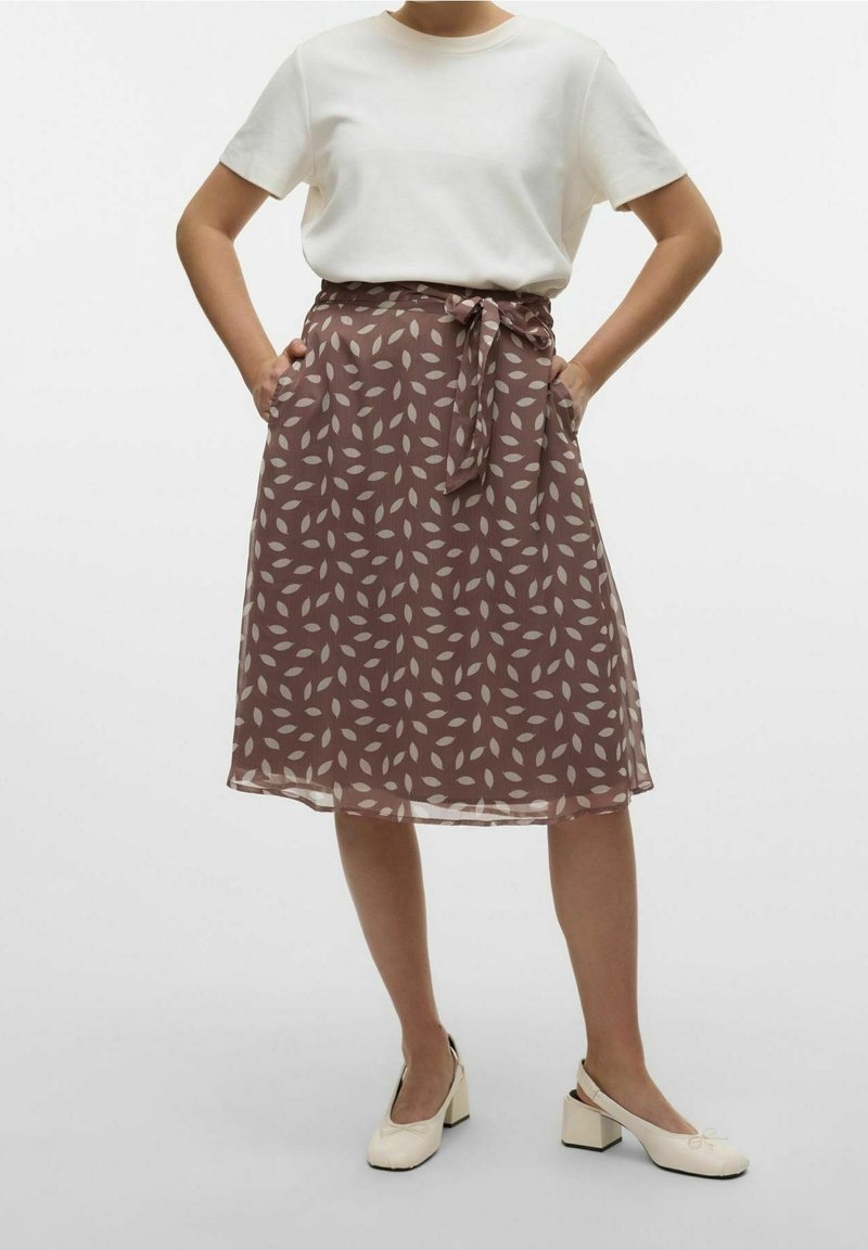 Femme portant un haut blanc à manches courtes, une jupe marron longueur genou avec un motif léger de feuilles et une ceinture nouée, ainsi que des chaussures à talons blocs de couleur crème.
