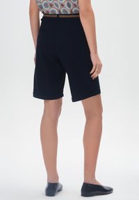 Czarne szorty do kolan o gładkiej fakturze, z mankietami, kieszeniami z tyłu i klasycznym wzornictwem. Noszone z płaskimi czarnymi butami.