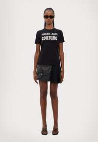 Must musta, millel on "Versace Jeans Couture" logo, koos musta mini-seelikuga, millel on taskud ja kuldne klamber. Mustad prillid.