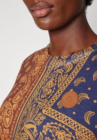 Top de manga corta con un patrón de paisley azul y óxido, con una etiqueta texturizada en marrón que muestra un logo de sol en la parte frontal. Escote circular.