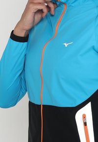 Blue and black athletic jacket with orange zipper and accents, featuring a zippered pocket and textured fabric in the lower portion.