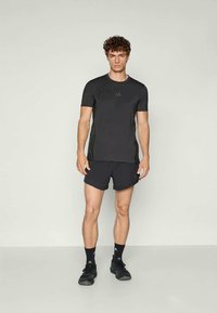 Tânăr stând în picioare, îmbrăcat cu un tricou sportiv negru, pantaloni scurți negri, ciorapi negri și pantofi sport negri, pe un fundal gri simplu.