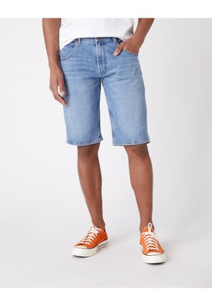 Homme portant un short en jean bleu clair, un t-shirt blanc et des baskets orange, debout avec une main dans la poche sur un fond blanc uni.