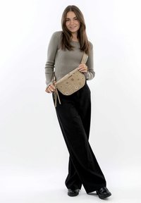 Beige crossbodytas met ronde uitsparingen, gemaakt van glad materiaal. Voorzien van verstelbare riem en kwastdetail. Gecombineerd met een grijze geribbelde trui.