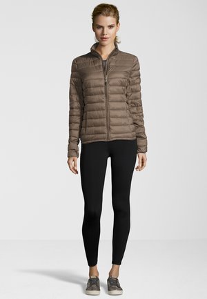 Bruine pufferjack met hoge kraag en ritssluiting, voorzien van een geborduurde textuur. Gecombineerd met zwarte legging en grijze sneakers.