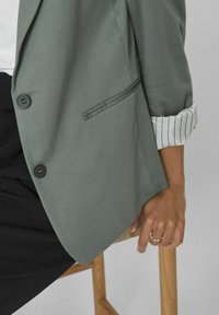 Osoba siedząca na drewnianym stołku, ubrana w szary żakiet z podwiniętymi paskami, czarne spodnie i srebrny pierścionek na lewej ręce.