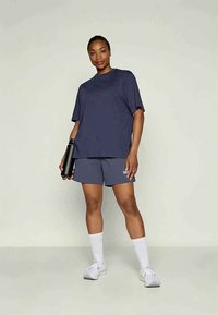 Sieviete tumši zila oversize T-kreklā un šortos, tur melnu ūdens pudeli, ģērbusies baltās kedās un zeķēs, stāvot pret vienkāršu gaišu fona.