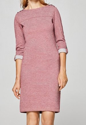 Femme portant une robe en maille rose, longueur genou, à manches longues avec revers roulés et détails de boutons sur les épaules et les manches.