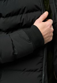 Crna jakna s punjenjem, s rebrastim manžetama, ima glatku vanjštinu i zatvaranje na patentni zatvarač, ističući ugodnu izolaciju i elegantan dizajn.