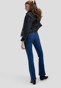 Žena pozira u tamnoj teksas jakni, plavim bootcut trapericama i crnim gležnjačama na jednostavnoj sivoj pozadini.