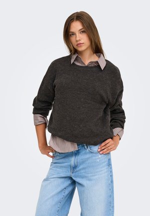 Jeune femme aux longs cheveux bruns portant un pull gris foncé par-dessus une chemise rayée et un jean bleu clair, debout les mains sur les hanches.