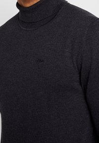 Mørkegrå rullekravesweater lavet af blødt strikstof; har en høj krave og diskret branding på brystet.