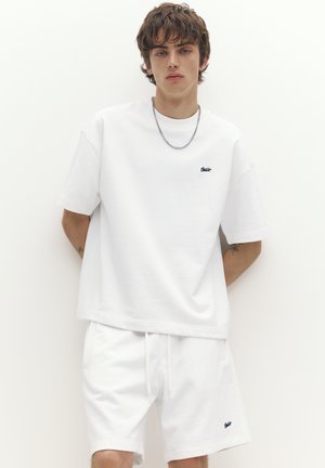 Junge Person trägt ein weißes übergroßes T-Shirt und passende weiße Shorts mit kleinen schwarzen Logos, steht vor einem schlichten weißen Hintergrund.