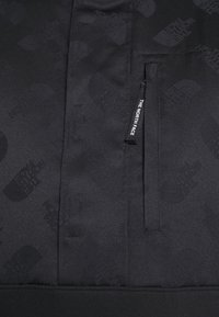 Svart tröja med slät textur och ett subtilt logomönster, horisontell dragkedjeficka och en texturerad halsringning. Överlag minimalistisk design.