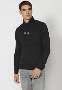 Črn pleten pulover z visokim ovratnikom in vrvicami, z teksturno zasnovo in logotipom. V kombinaciji s svetlo sivo jeans materialom.