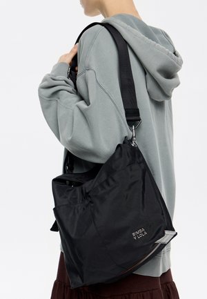 Czarna nylonowa torebka o teksturowanej powierzchni, wyposażona w dużą przednią kieszeń oraz regulowany pasek. Logo marki widoczne z boku.