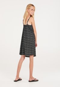 Robe sans manches noire avec des motifs géométriques blancs, silhouette évasée, longueur mi-cuisse, et bretelles réglables ; portée avec des tongs noires.