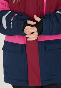 Farverig pufferjakke med maroon, pink og navy sektioner. Vandafvisende materiale med reflekterende detaljer og justerbare manchetter.
