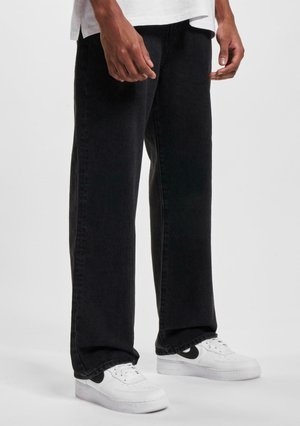 Jean droit noir en denim avec une texture lisse, assorti à des baskets blanches avec des accents noirs. Design simple, sans motifs visibles.