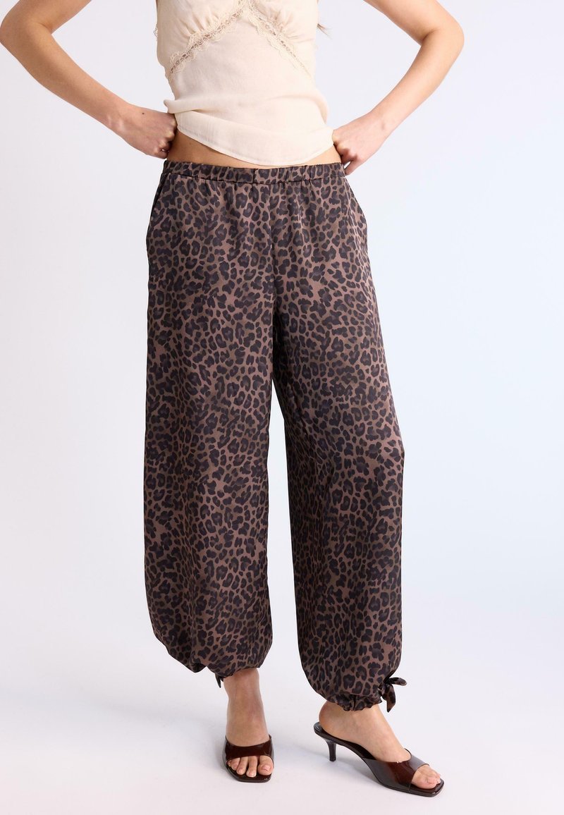 Femme portant un pantalon ample à imprimé léopard marron avec élastique et nœuds aux chevilles, associé à un haut beige orné de dentelle et des sandales à talons foncées.
