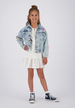 Lichtblauwe spijkerjasje met kleurrijke patches, gecombineerd met een witte eyelet jurk met een gerimpelde zoom, en marineblauwe hoge sneakers.