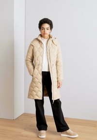 Abrigo largo acolchado de un beige claro con capucha, cierre frontal, cintura con cordón y bolsillos laterales, llevado sobre un suéter de punto blanco y pantalones negros.