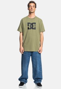 Camiseta de algodón color verde oliva con un gran logo negro de DC en la parte frontal, combinada con jeans holgados de color azul y zapatillas negras.