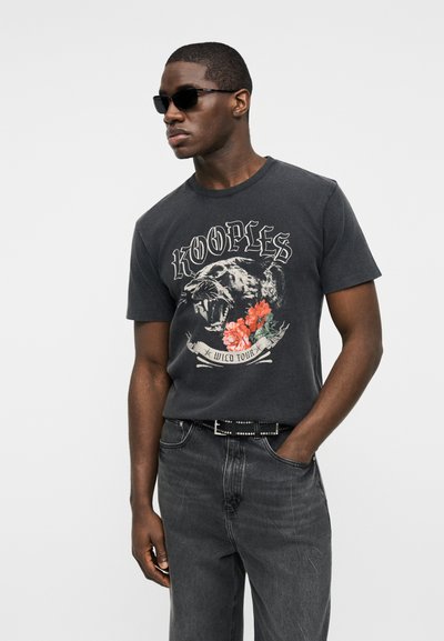 Άνδρας που φοράει μαύρα γυαλιά ηλίου, μια μαύρη μπλούζα με γραφικό σχέδιο ενός ουρλιάζοντος πάνθηρα και λουλούδια, και σκούρα γκρι τζιν με ζώνη διακοσμημένη με καρφιά.