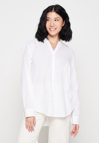 Camicia bianca con colletto a V, maniche lunghe e una vestibilità rilassata. Il tessuto appare liscio e leggero. I bottoni sono di colore chiaro.