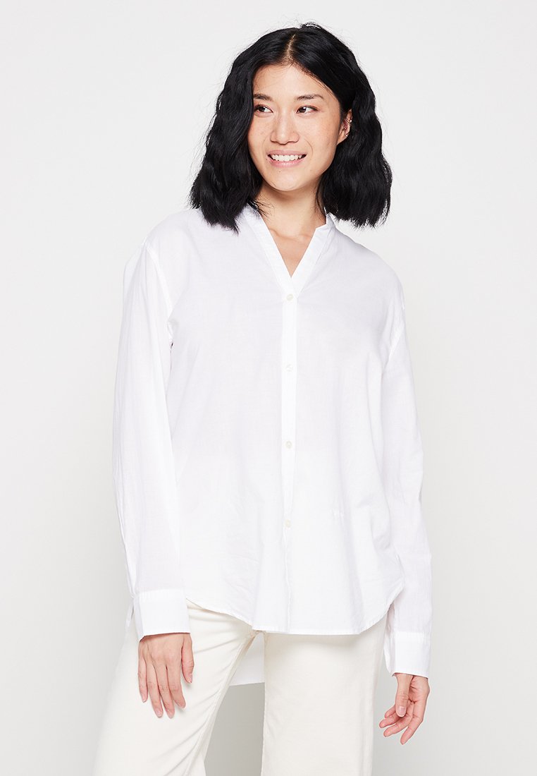 Camicia bianca con colletto a V, maniche lunghe e una vestibilità rilassata. Il tessuto appare liscio e leggero. I bottoni sono di colore chiaro.