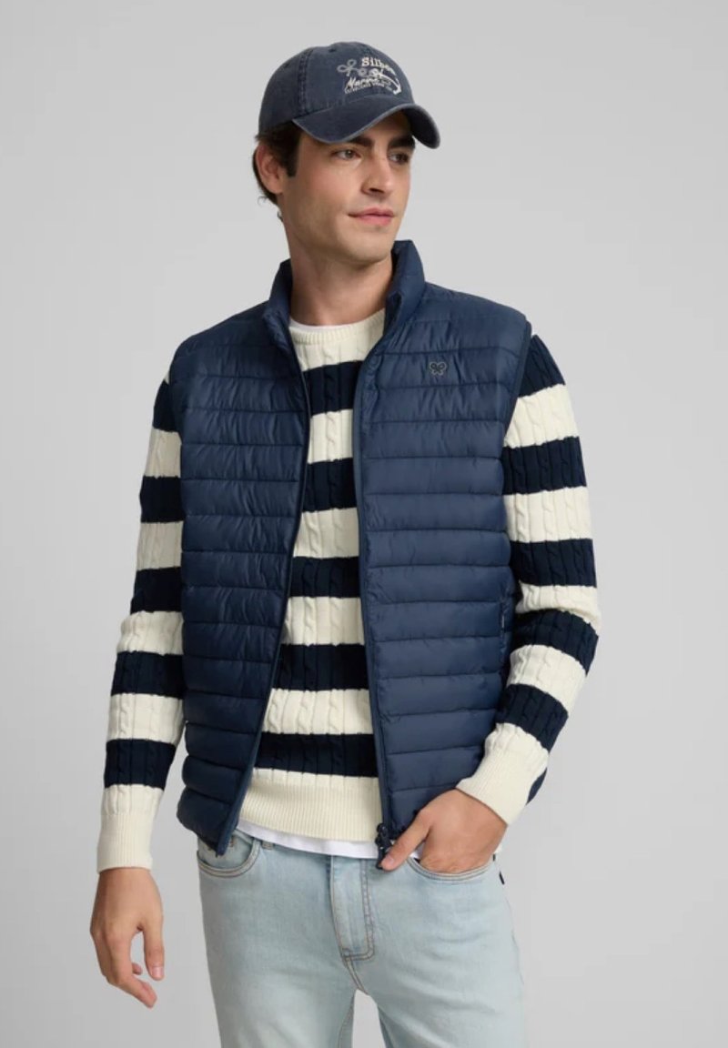 Chaleco acolchado azul marino con cierre frontal, llevado sobre un suéter de punto de rayas crema y azul marino. El modelo lleva una gorra azul marino a juego.