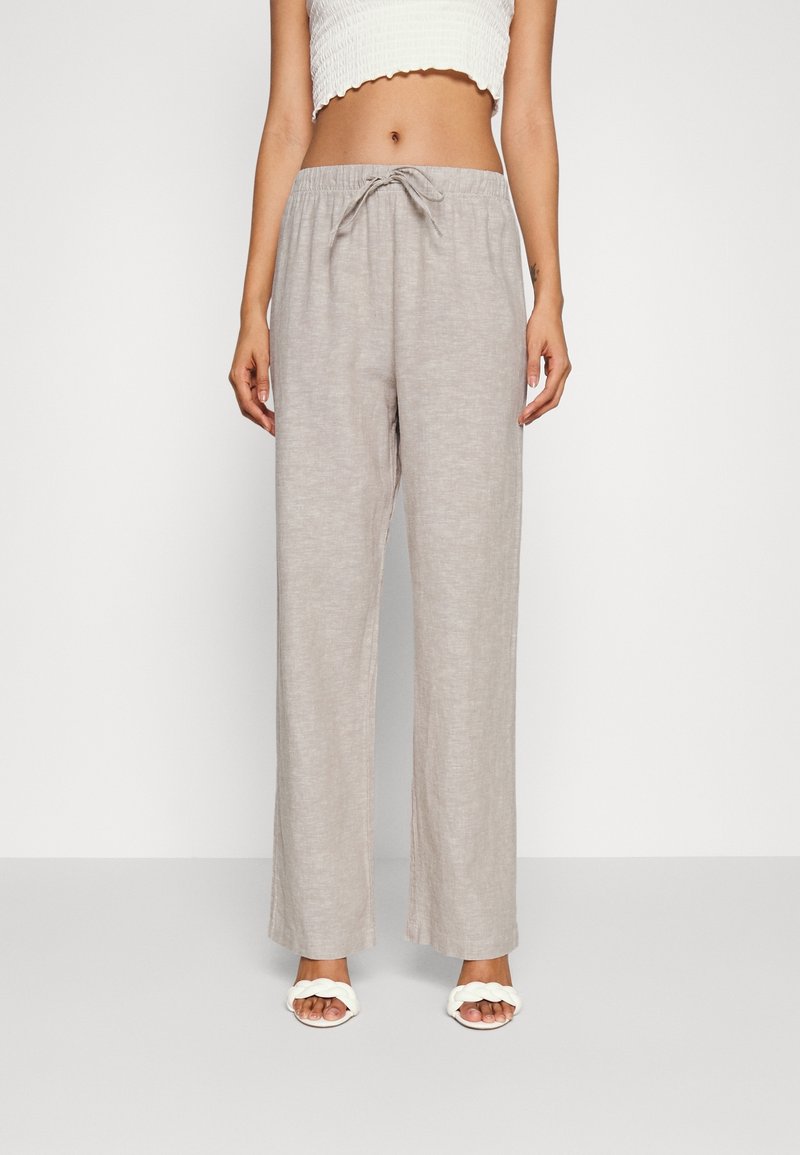 Femme portant un pantalon ample gris clair à cordon, un top court blanc et des sandales blanches tressées, debout devant un fond uni.