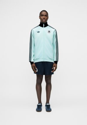 Vyras stovintis, dėvintis šviesiai mėlyną Adidas sportinę striukę, juodus šortus ir mėlynas sportbačius prieš paprastą pilką foną.