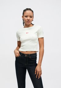 Valkoinen ribbattu lyhyt paita, jossa on lyhyet hihat ja punainen sydänkuvio rinnassa. Yhdistetty tummiin farkkuihin. Sileä tekstuuri.