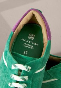 Baskets en daim vert avec des lacets blancs et verts, doublure intérieure rembourrée beige et violette, et semelle intérieure verte marquée "Signature Forever Comfort".