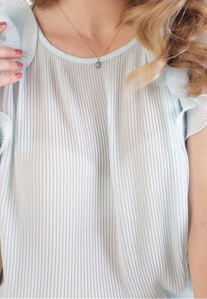 Femme portant un chemisier sans manches plissé bleu clair avec des bords volantés et un petit pendentif, main levée près de l'épaule.