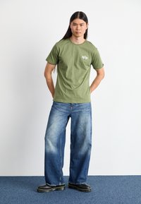 Kortærmet T-shirt i olivengrøn med afslappet pasform og hvidt logo. Kombineret med blå vidde-bukser og sorte platform-støvler. Minimalistisk design.