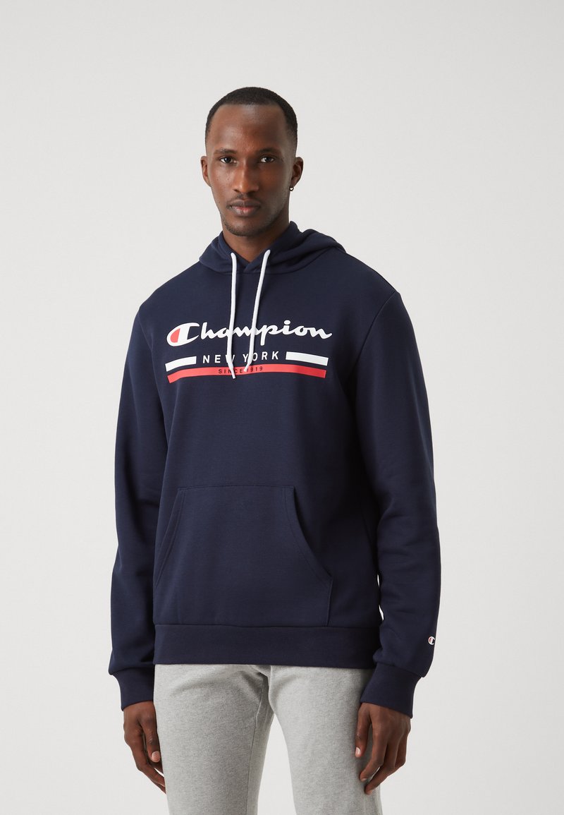 Tumši zils kapuci krekls ar baltām saites, priekšējo kabatu un drukātu "Champion New York" logo baltās, sarkanās un melnās svītrās.
