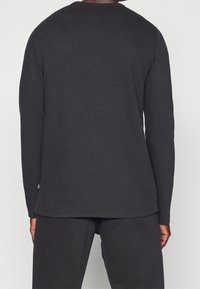 Schwarz langärmliges Thermoshirt aus strukturiertem Stoff mit rundem Ausschnitt; hat einen geraden Saum und ein dezentes Logo-Detail am Saum.