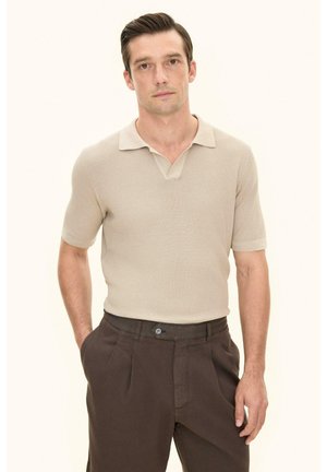 Mand med kort mørkt hår iført en beige kortærmet strikket polo og mørkebrune bukser, stående med den ene hånd i lommen.