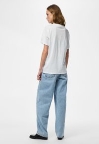 Femeie stând pe o parte, purtând un tricou simplu alb cu mâneci scurte, blugi largi albastru deschis și pantofi negri pe un fundal alb simplu.