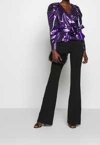 Haut violet brillant à manches longues avec épaules volumineuses, taille cintrée et un nœud. Associé à un pantalon noir évasé et une pochette noire texturée.