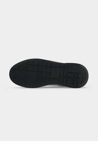 Semelle de chaussure noire avec un motif de bande de roulement en forme de grille, profil plat et surface texturée pour la traction, présentée sur un fond clair uni.