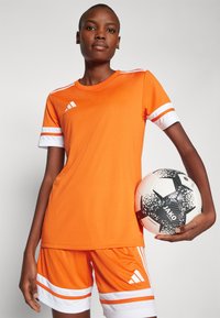 Oranje sportshirt met witte strepen op de mouwen, gecombineerd met bijpassende shorts. Model houdt een zwart-witte voetbal vast.
